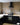 A black kitchen with white work surfaces, a black cooker hood is backed by yellow brick slips on the wall behind.