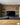 A TV, and houseplants are infront of a natural creased brown brick slip wall, with a wooden table in the foreground.